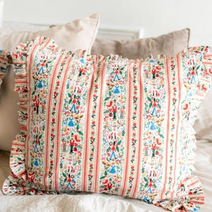 May include: A square throw pillow with a ruffled edge featuring a repeating vertical stripe pattern. The design includes colorful illustrations of nutcrackers, dancers, Christmas stockings, gifts, and floral motifs on a white background.