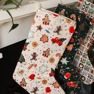 May include: Christmas stockings with festive patterns. One stocking features gingerbread men, houses, and trees on a cream background. Other stockings have green and red patterns. Each stocking has a loop for hanging.