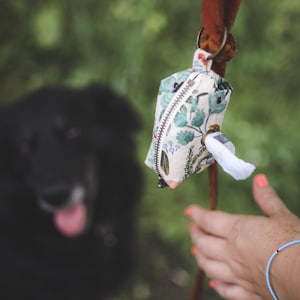 May include: A floral patterned dog waste bag dispenser with a white tissue inside, attached to a brown cord. A black dog is out of focus in the background.