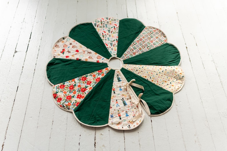 May include: A circular, scalloped-edge tree skirt featuring alternating panels of solid dark green fabric and patterned fabric. The patterns include nutcracker figures, red poinsettias, and festive holiday motifs. It has a center opening and ties.