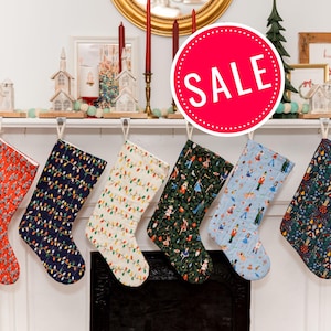 May include: Six Christmas stockings with various patterns, including red with white designs, navy with string lights, and a light blue stocking with figures. The stockings hang from a white mantelpiece, with a red "SALE" sign in the background.