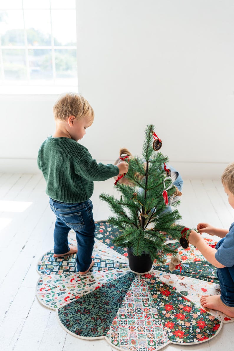 May include: A circular fabric Christmas tree skirt featuring a patchwork design of various holiday-themed patterns including floral motifs, holly leaves, and festive icons in shades of red, green, white, and dark blue with a scalloped edge.