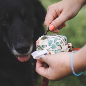 May include: A person holding a small floral patterned bag with a zip. The bag is attached to a brown leather lead. A black dog is in the background.