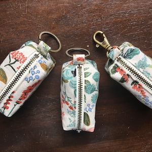 May include: Three small zippered pouches with floral patterns and gold-toned key rings. The pouches are made of fabric and have a white background with colourful flowers and leaves.