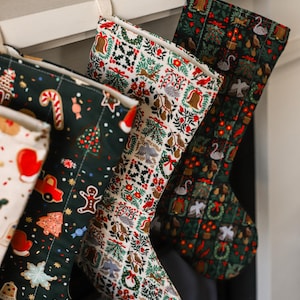 May include: Three Christmas stockings hanging in a row. The left stocking is dark green with gingerbread men, candy canes, and red trucks. The middle stocking is cream with a grid of holiday motifs. The right stocking is dark green with swans.