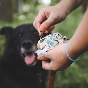 May include: A person's hand holding a small floral patterned bag with a zip. The bag is attached to a brown lead. A black dog is in the background.