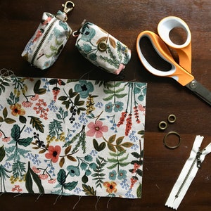 May include: A close-up of a fabric project with a floral pattern. The project includes two small pouches with a zipper and a key ring, a pair of scissors, two metal rings, and a zipper.