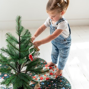 May include: A circular Christmas tree skirt featuring a patchwork design of various holiday-themed fabrics, including patterns of poinsettias, gingerbread cookies, candy canes, and evergreen trees, placed under a small artificial pine tree.
