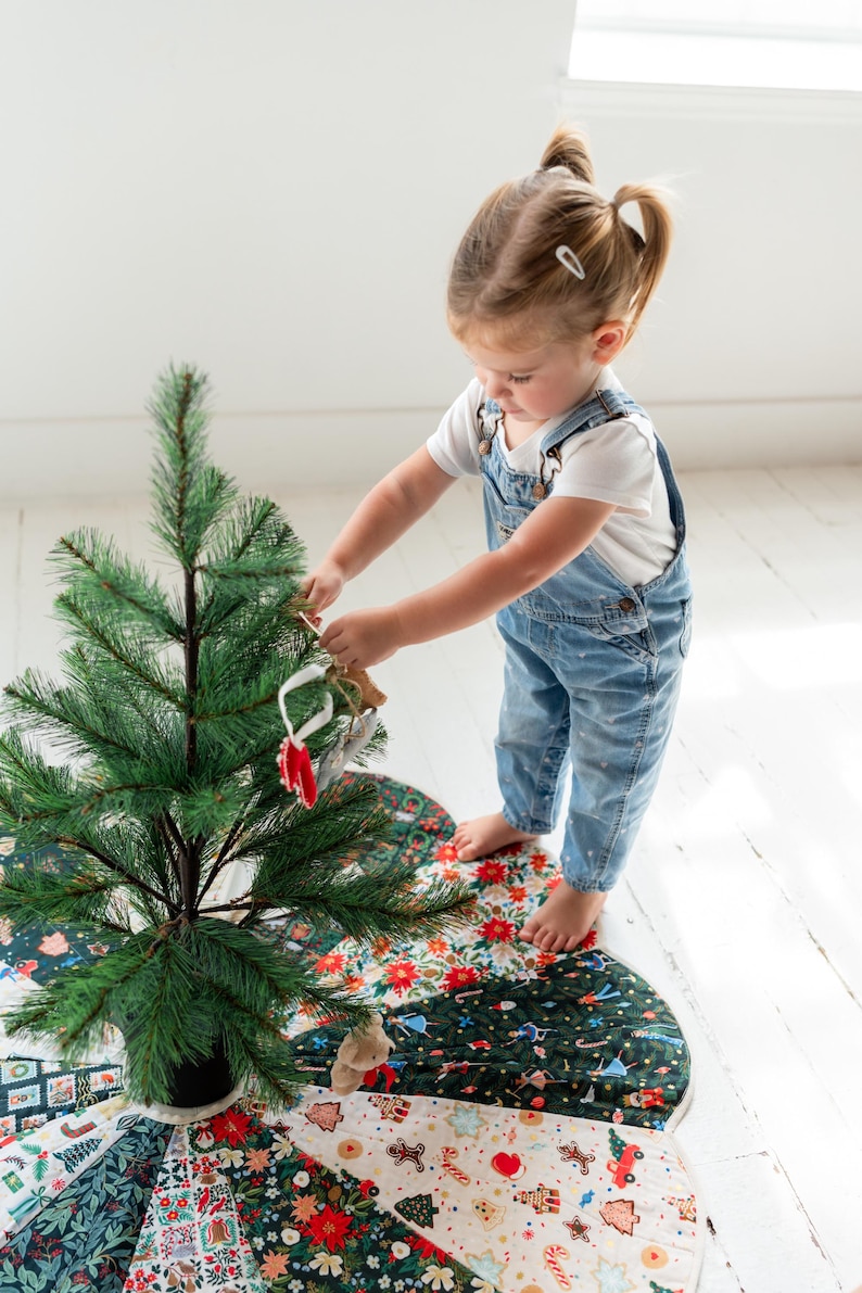 May include: A circular Christmas tree skirt featuring a patchwork design of various holiday-themed fabrics, including patterns of poinsettias, gingerbread cookies, candy canes, and evergreen trees, placed under a small artificial pine tree.