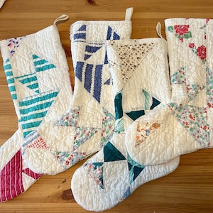 May include: Four quilted Christmas stockings featuring various patchwork patterns in white, teal, blue, pink, and floral prints. Each stocking has a fabric loop at the top for hanging and is constructed from stitched fabric panels with a textured finish.