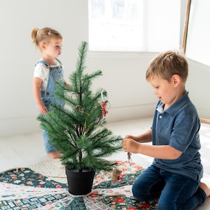 May include: A circular fabric tree skirt featuring a patchwork design of various holiday-themed patterns in shades of green, red, and white. The skirt has a scalloped edge and is placed under a small artificial green Christmas tree.