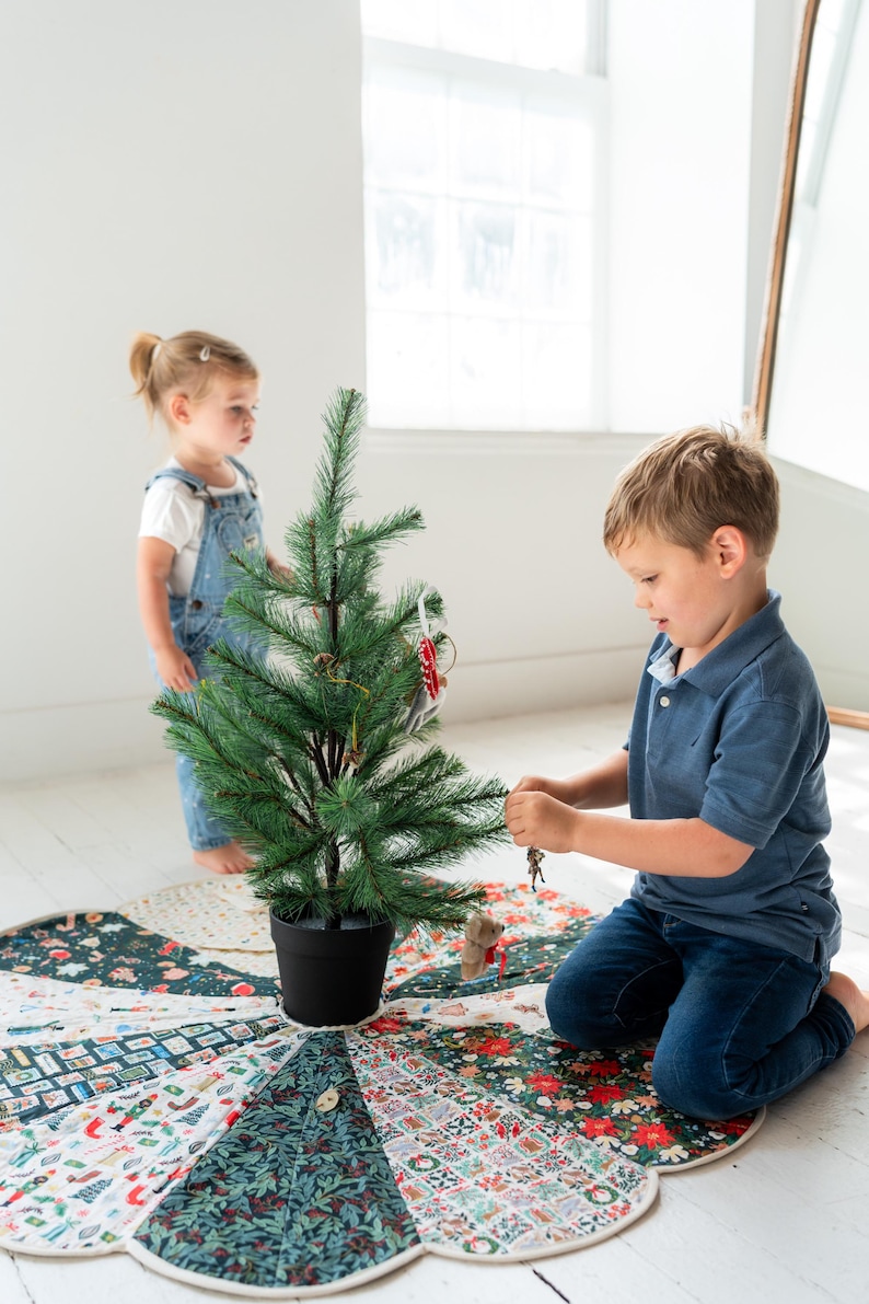 May include: A circular fabric tree skirt featuring a patchwork design of various holiday-themed patterns in shades of green, red, and white. The skirt has a scalloped edge and is placed under a small artificial green Christmas tree.