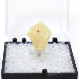 Może przedstawiać: Jasnożółty, krystaliczny okaz minerału w przezroczystym plastikowym etui z czarną podstawą. Minerał jest zamontowany na przezroczystym stojaku i otoczony małymi, białymi kawałkami przypominającymi piankę. Etui jest otwarte.
