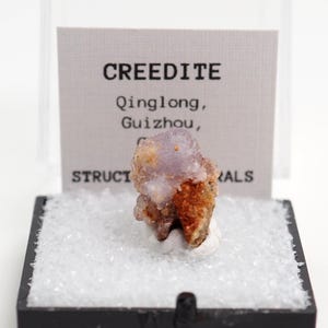 May include: A Creedite mineral specimen from Qinglong, Guizhou, China, presented in a clear display case. The mineral features a mix of purple, brown, and white hues. The display case includes a label with the mineral's name and origin.