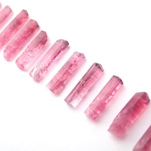 Puede incluir: Disposición diagonal de varitas de cristal de turmalina rosa sobre fondo blanco. Los cristales, de tamaños y formas variadas, presentan un aspecto translúcido y tonos rosados.