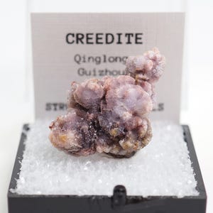 May include: A Creedite mineral specimen from Qinglong, Guizhou, China, presented in a black display case. The mineral has a clustered, crystalline structure with a pale purple hue and a touch of brown.