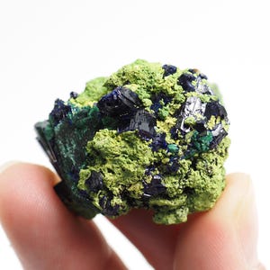 Könnte beinhalten: Nahaufnahme einer Mineralprobe mit einer Farbkombination. Das Mineral hat eine leuchtend grüne Basis mit dunkelblauen und grünen kristallinen Strukturen. Die Probe wird vor einem weißen Hintergrund gehalten.