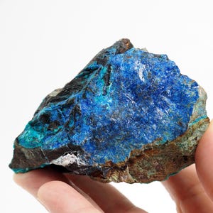 Könnte beinhalten: Ein rohes Mineralexemplar mit tiefblauen und türkisfarbenen Farbtönen. Die Oberfläche des Gesteins ist strukturiert, mit Bereichen in Dunkelbraun und Schwarz. Das Mineral wird in einer Hand gehalten, was seine natürliche Schönheit hervorhebt.