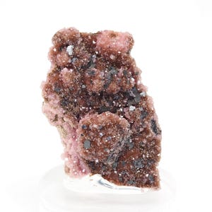 May include: A mineral specimen of rhodochrosite, displaying a cluster of pink crystals with dark grey and black inclusions. The mineral sits on a clear base against a white backdrop. The color scheme is a blend of pink, brown, and grey.