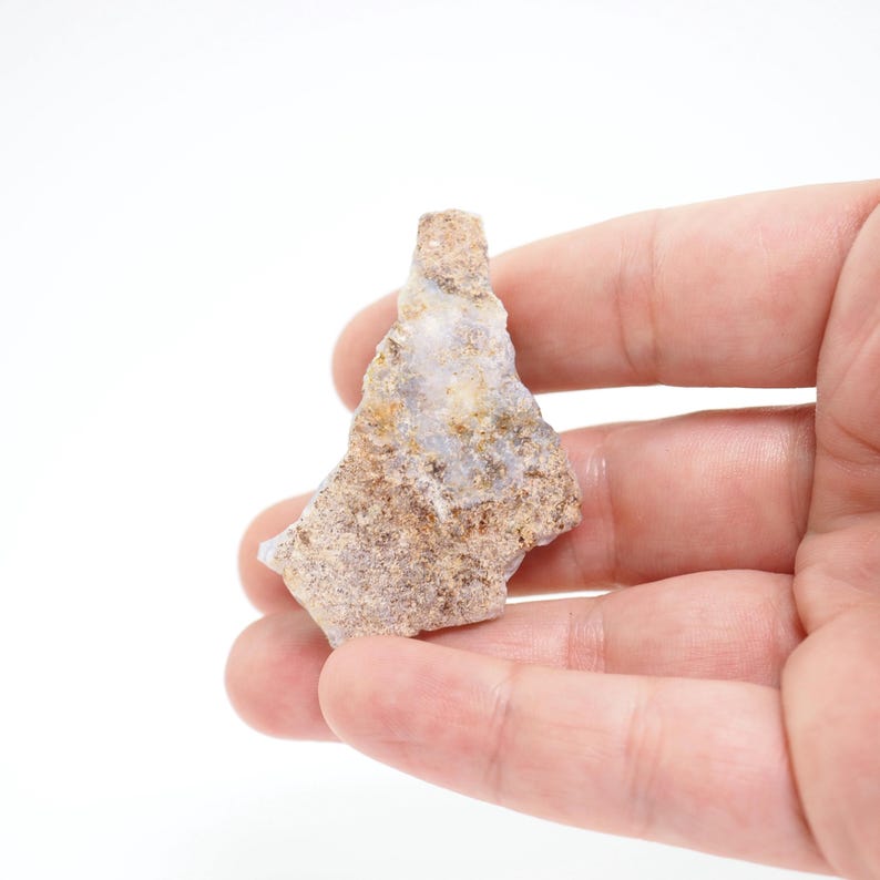 May include: A rough, irregularly shaped stone with a mix of tan, brown, and pale blue hues. The stone has a textured surface and is held in a hand against a white background. The stone appears to be a natural mineral specimen.
