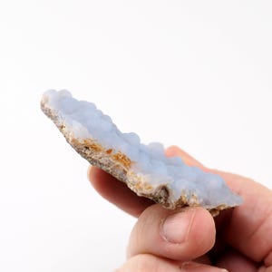 Puede incluir: Una pieza de piedra preciosa de calcedonia azul con una textura burbujeante. La piedra tiene un color azul claro con un borde &aacute;spero y marr&oacute;n. La gema se sostiene en una mano sobre un fondo blanco.
