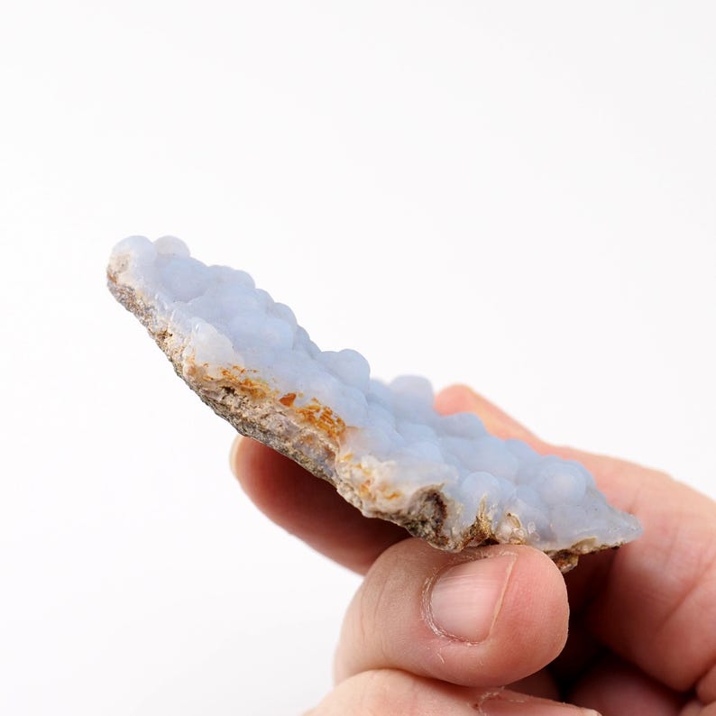 Puede incluir: Una pieza de piedra preciosa de calcedonia azul con una textura burbujeante. La piedra tiene un color azul claro con un borde &aacute;spero y marr&oacute;n. La gema se sostiene en una mano sobre un fondo blanco.