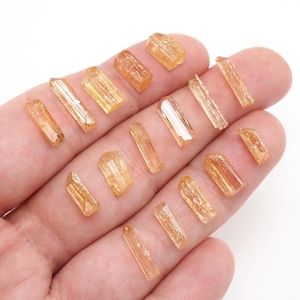 May include: A collection of small, translucent, golden-orange crystal shards with elongated, prismatic shapes and faceted surfaces. These natural mineral specimens exhibit varying lengths and widths, displaying a warm, honey-toned clarity throughout.