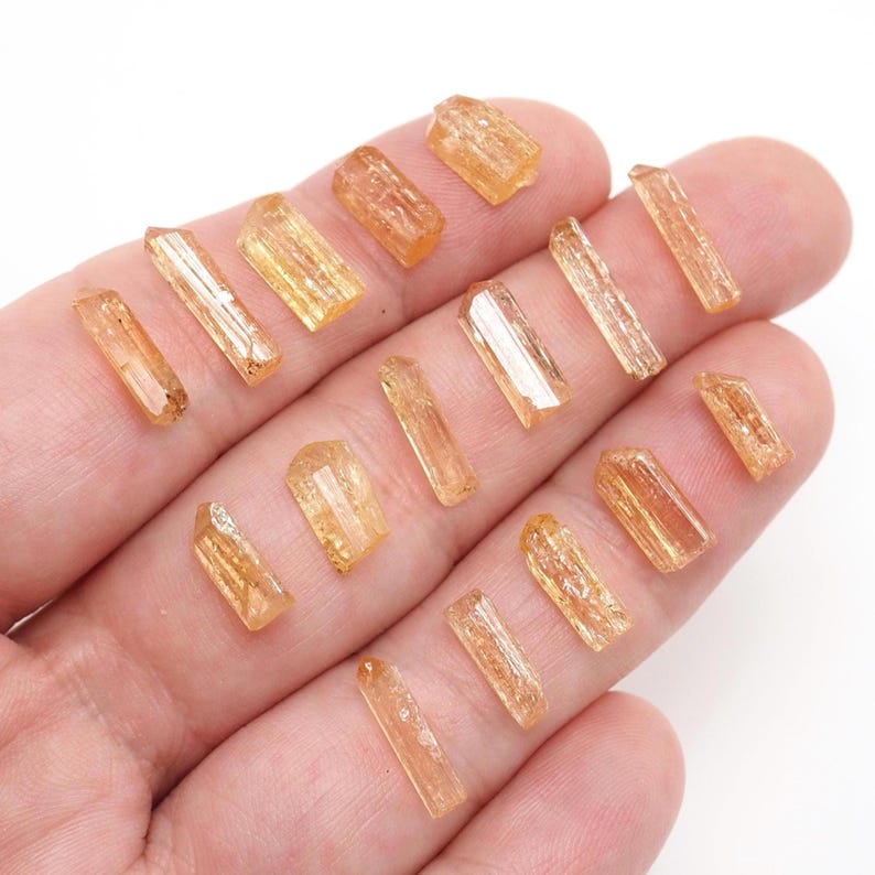 May include: A collection of small, translucent, golden-orange crystal shards with elongated, prismatic shapes and faceted surfaces. These natural mineral specimens exhibit varying lengths and widths, displaying a warm, honey-toned clarity throughout.