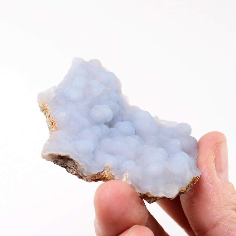Puede incluir: Un esp&eacute;cimen de calcedonia azul claro con una textura botrioidal y burbujeante. La piedra tiene un borde &aacute;spero y marr&oacute;n y se sostiene en una mano sobre un fondo blanco. Este cristal natural es una pieza &uacute;nica para coleccionistas.