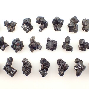 Puede incluir: Una colecci&oacute;n de 18 cristales negros, algunos con matices iridiscentes verdes y azules, dispuestos en filas sobre un fondo blanco.