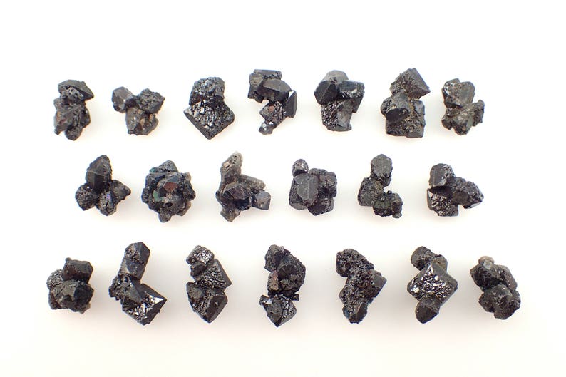 Puede incluir: Una colecci&oacute;n de 18 cristales negros, algunos con matices iridiscentes verdes y azules, dispuestos en filas sobre un fondo blanco.