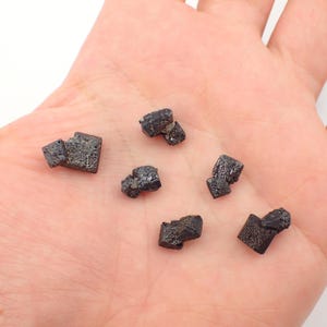 Puede incluir: Una colecci&oacute;n de seis peque&ntilde;os cristales negros, formados naturalmente, con una superficie rugosa e irregular. Los cristales est&aacute;n dispuestos en una mano.