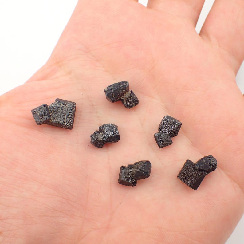 Puede incluir: Una colecci&oacute;n de seis peque&ntilde;os cristales negros, formados naturalmente, con una superficie rugosa e irregular. Los cristales est&aacute;n dispuestos en una mano.