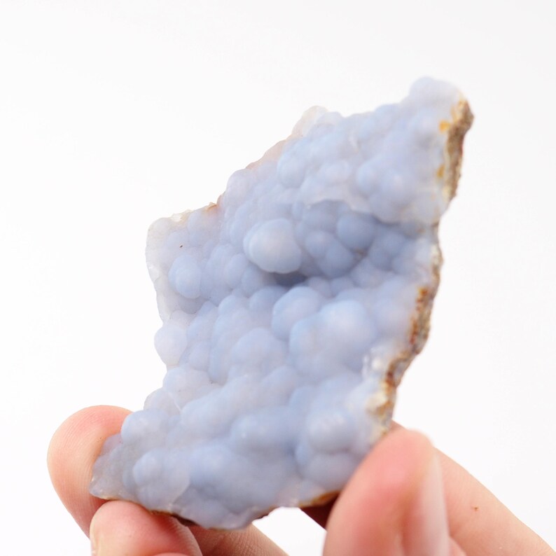 Puede incluir: Una pieza de piedra preciosa de calcedonia azul con una superficie texturizada y burbujeante. La piedra se sostiene en una mano sobre un fondo blanco. Los bordes de la piedra tienen un tono marr&oacute;n y terroso.