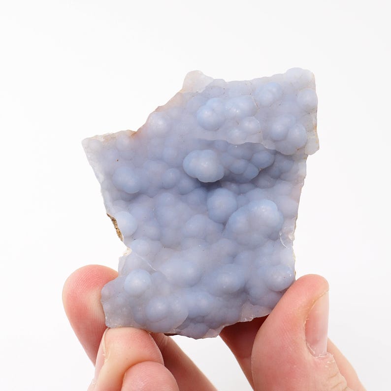 Puede incluir: Muestra de mineral de calcedonia azul claro con textura botrioidal. La piedra, sobre un fondo blanco, muestra formaciones redondeadas y un tono p&aacute;lido. El borde natural del mineral es visible, resaltando su forma bruta.