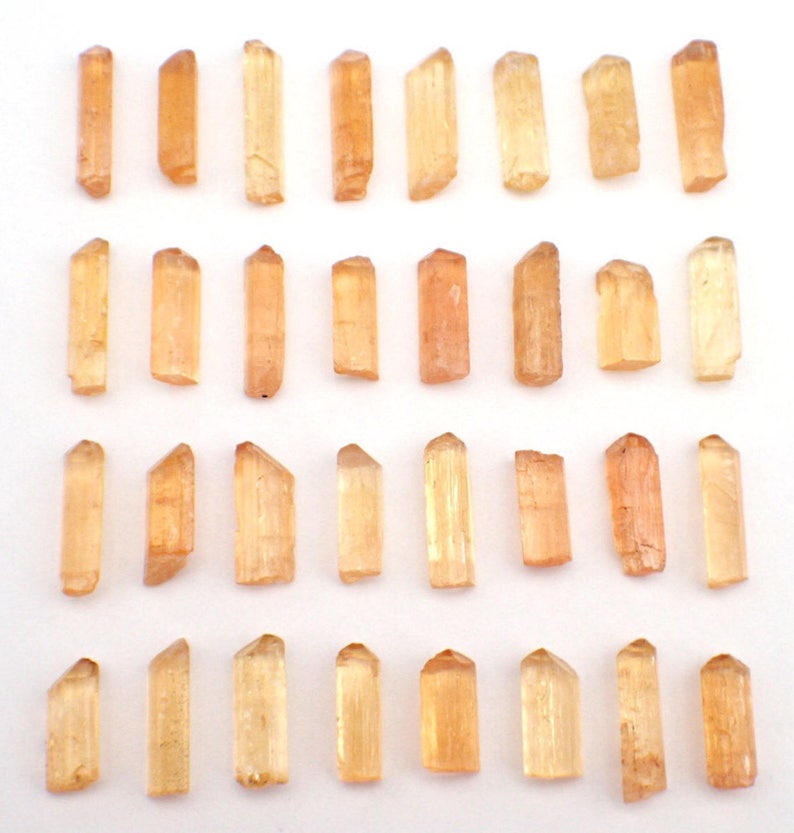 May include: A collection of thirty-two small, translucent, light orange crystal points arranged in four horizontal rows. Each individual crystal features a prismatic shape with a pointed termination and a smooth, elongated, rectangular body.