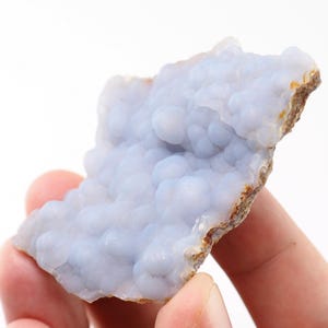 Puede incluir: Una pieza de piedra preciosa de calcedonia azul claro con una textura botrioidal y burbujeante. La piedra tiene un borde &aacute;spero y marr&oacute;n y se sostiene en una mano sobre un fondo blanco. El color azul es suave y pastel.