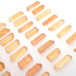 May include: A collection of numerous small, elongated, translucent crystals in shades of light orange and golden yellow. Each crystal features a natural, prismatic shape with pointed or faceted ends, arranged in a loose, scattered pattern.