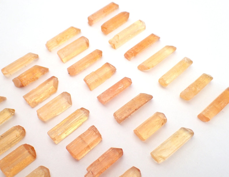 May include: A collection of numerous small, elongated, translucent crystals in shades of light orange and golden yellow. Each crystal features a natural, prismatic shape with pointed or faceted ends, arranged in a loose, scattered pattern.