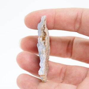 May include: A light blue and brown raw gemstone slice with a textured surface. The stone is held in a hand, showcasing its natural, irregular shape and color variations. The gemstone has a rough, earthy appearance.
