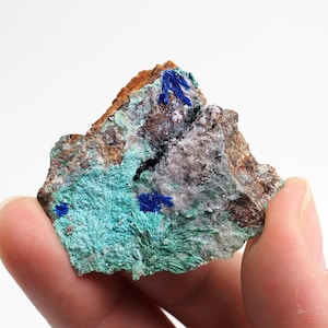 May include: A rough-hewn rock sample featuring a mix of colors and textures. The rock displays areas of brown, gray, and turquoise, with vibrant blue crystalline formations. The specimen is held against a white background.