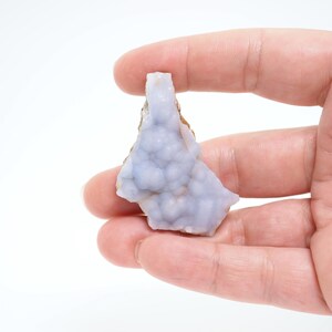 May include: A light blue, natural mineral specimen with a bubbly texture. The stone is held in a hand against a white background. The mineral has a rough, brown edge and a smooth, rounded surface.