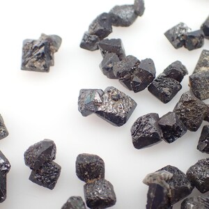 Puede incluir: Una colecci&oacute;n de peque&ntilde;os cristales octa&eacute;dricos negros. Los cristales est&aacute;n dispuestos sobre una superficie blanca.