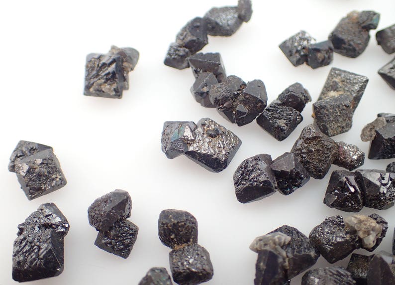Puede incluir: Una colecci&oacute;n de peque&ntilde;os cristales octa&eacute;dricos negros. Los cristales est&aacute;n dispuestos sobre una superficie blanca.