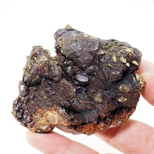 Puede incluir: Un espécimen mineral en bruto sin cortar, con un grupo de estructuras cristalinas de color rojo oscuro. El mineral tiene una superficie rugosa y texturizada con parches de inclusiones metálicas doradas. La paleta de colores es una mezcla de rojos oscuros, marrones y dorados.