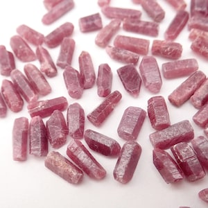 May include: A collection of small, pink, raw ruby crystals in a variety of shapes and sizes. The crystals are scattered on a white surface.