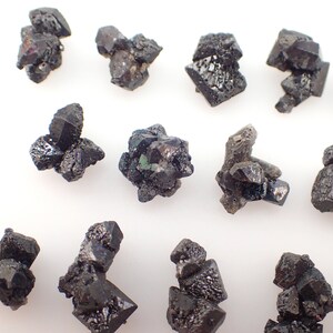 Puede incluir: Una colecci&oacute;n de cristales negros y verdes, algunos con un brillo met&aacute;lico, dispuestos sobre una superficie blanca. Los cristales son de varios tama&ntilde;os y formas, algunos parecen m&aacute;s puntiagudos que otros.
