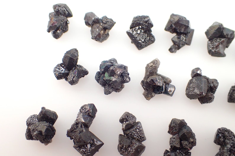 Puede incluir: Una colecci&oacute;n de cristales negros y verdes, algunos con un brillo met&aacute;lico, dispuestos sobre una superficie blanca. Los cristales son de varios tama&ntilde;os y formas, algunos parecen m&aacute;s puntiagudos que otros.