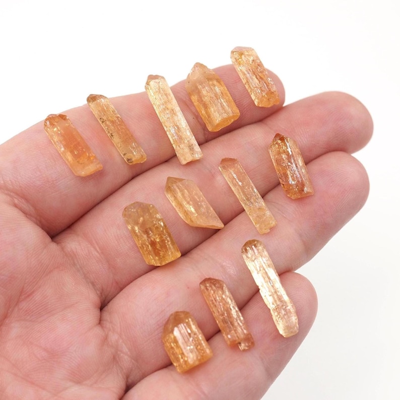 May include: A collection of eleven small, translucent, orange-hued crystal points with elongated, prismatic shapes and natural facets. These individual mineral specimens exhibit varying lengths and widths, displaying a warm, golden-amber color throughout.