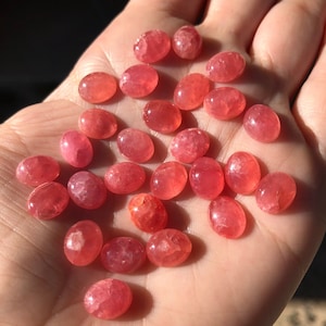 K&ouml;nnte beinhalten: Eine Sammlung von 25 ovalen, rosafarbenen und roten Edelstein-Cabochons. Die Cabochons sind glatt und poliert, mit einem leichten Schimmer.